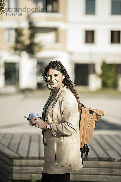 Junge Frau trägt Rucksack  hält Smartphone in der Hand und trinkt  während sie in der Stadt steht