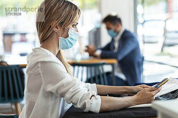 Nachdenkliche junge Frau trägt Schutzmaske  während sie während des Coronavirus-Ausbruchs im Café sitzt