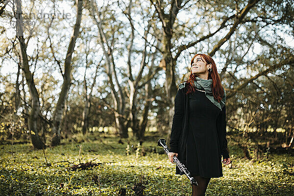 Junge lächelnde Frau hält Klarinette  blickt nach oben und geht im Wald spazieren
