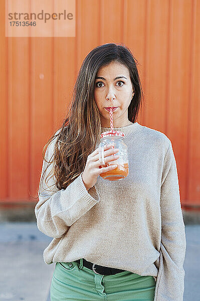 Schöne Frau trinkt Saft  während sie vor einer orangefarbenen Wand steht
