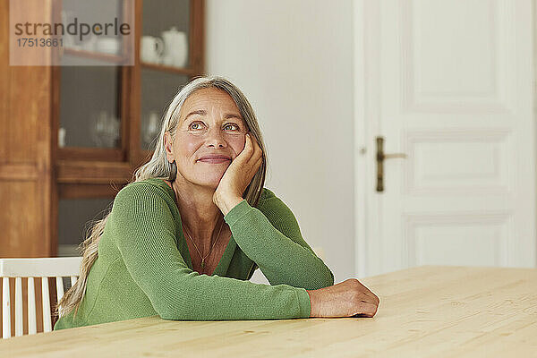 Lächelnde Frau träumt  während sie zu Hause am Tisch sitzt