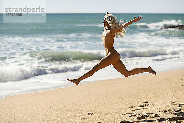 Sorglose nackte junge Frau springt am Strand gegen das Meer