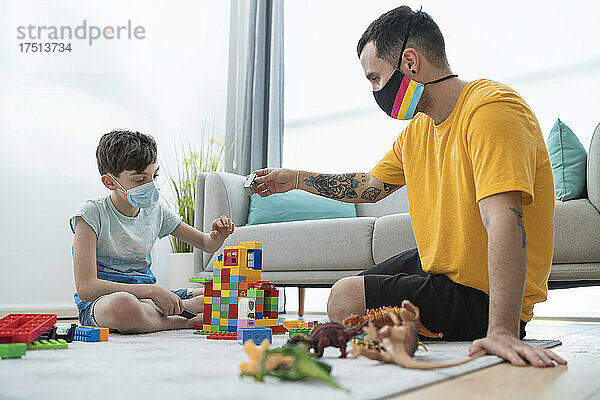 Mann und Junge tragen Masken  während sie während der Ausgangssperre von Covid-19 im Wohnzimmer mit Spielzeugklötzen spielen