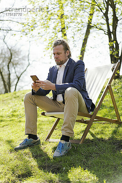 Reifer Geschäftsmann schreibt Textnachrichten per Mobiltelefon  während er auf einem Stuhl im Park sitzt