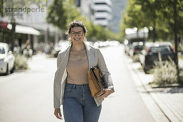 Lächelnde junge Frau trägt eine Tasche  während sie an einem sonnigen Tag auf der Straße läuft