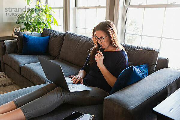 Frau sitzt auf Sofa im Wohnzimmer und arbeitet am Computer.