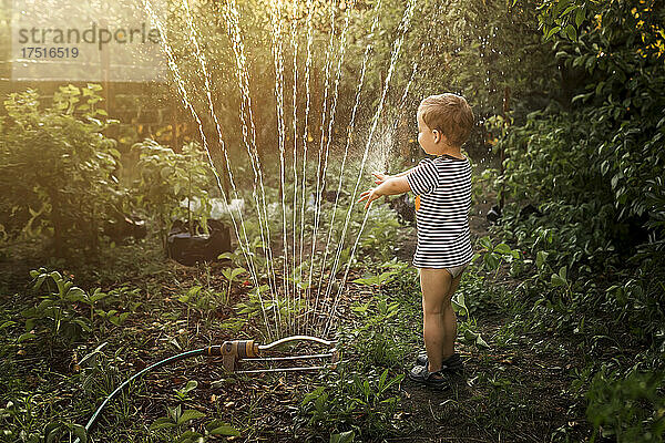 Seitenansicht eines kleinen blonden Jungen im Garten  der mit einem Wasserschlauch spielt