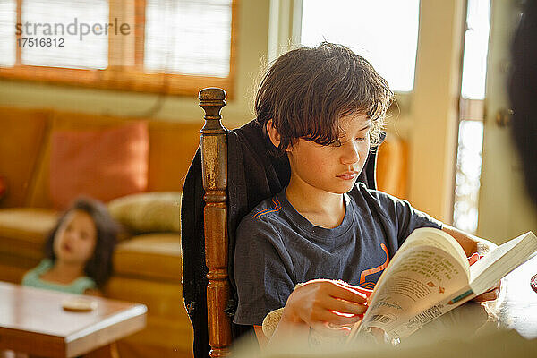 Ein Junge liest im Morgenlicht ein Buch  während sich seine Schwester hinter ihm entspannt