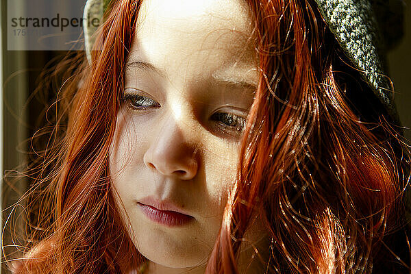 Ein ernstes Kind mit leuchtend roten Haaren steht im Fensterlicht