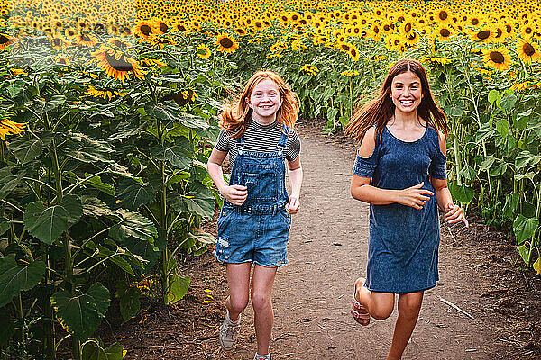 Zwei glückliche Tween-Mädchen laufen in einem Sonnenblumenfeld.