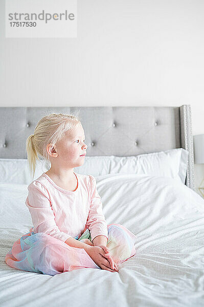 Junges Mädchen im rosa Regenbogenkleid sitzt auf einem Bett und schaut aus dem Fenster