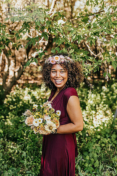 Lächelnde Frau steht mit Blumenstrauß und Blumenkrone im Wald