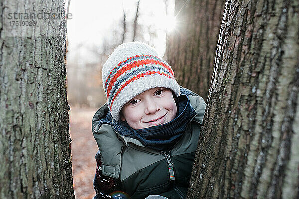 Junge blickt lächelnd durch Bäume im Winter bei Sonnenuntergang