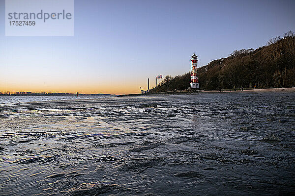 Eisiger Winternachmittag am Strand von Wittenbergen in Hamburg-Rissen. Die untergehende Sonne beleuchtet den Leuchtturm. Im Hintergrund die Schornsteine des Kohlekraftwerkes Wedel