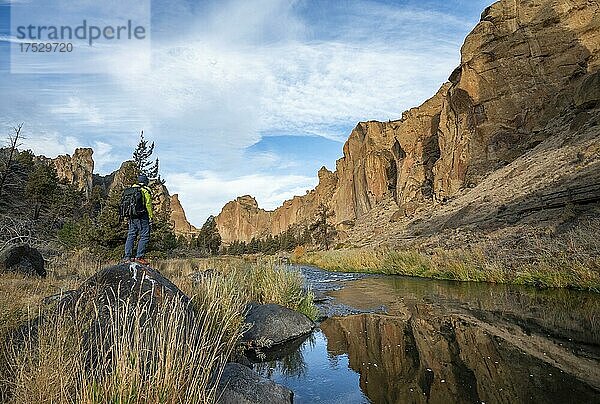 Junger Mann steht auf Stein am Ufer  rote Felswände in der Morgensonne  spiegeln sich im Fluss  Flusslauf des Crooked River  Canyon mit Felsformationen  The Red Wall  Smith Rock State Park  Oregon  USA  Nordamerika