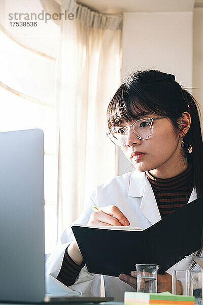 Scientist writing in book while looking at laptop at laboratory