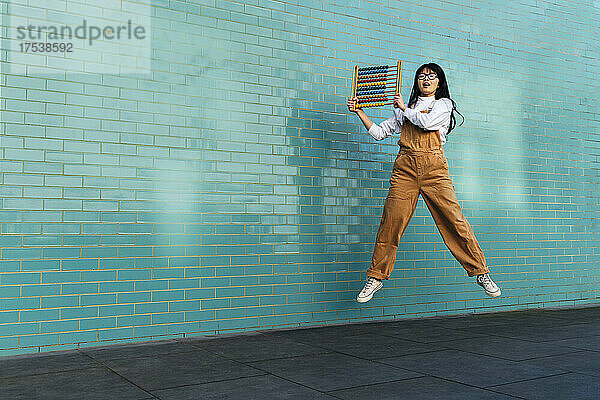 Cheerful woman holding abacus jumping in front of brick wall