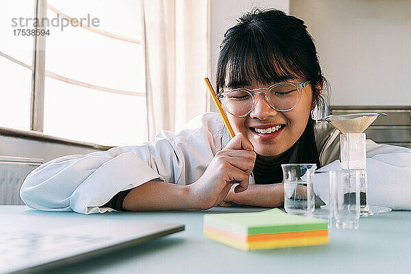 Happy scientist with pencil leaning on table in laboratory