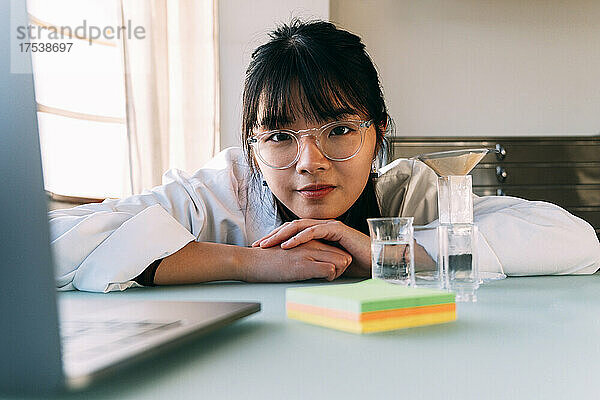Young chemist wearing eyeglasses leaning on table in laboratory
