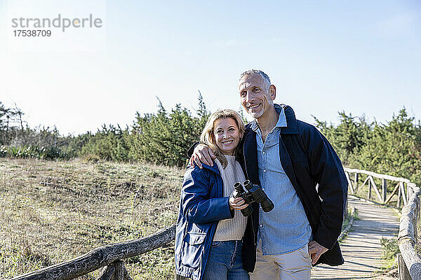 Man with arm around woman holding binoculars on wooden bridge