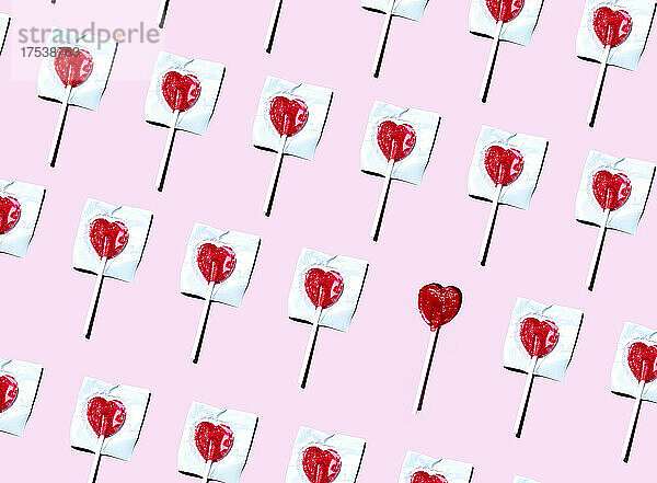 Pattern of heart shaped lollipops flat laid against pink background with single one unwrapped
