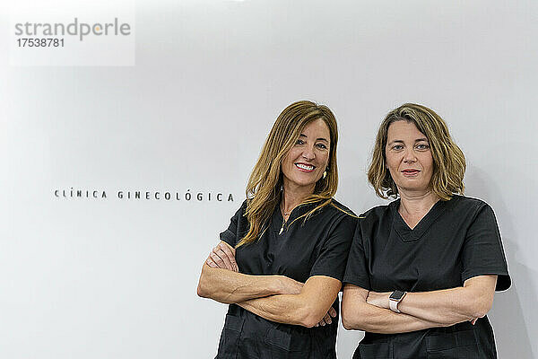 Confident female doctors standing with arms crossed in front of white wall in clinic
