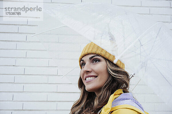 Lächelnde Frau mit transparentem Regenschirm an der Wand