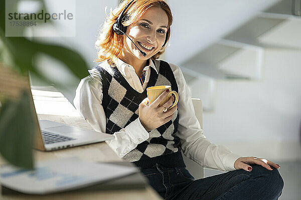 Lächelnde Geschäftsfrau mit Becher sitzt im Heimbüro