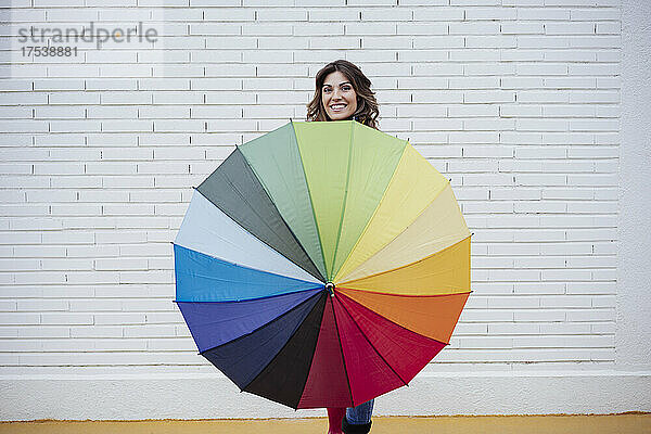 Smiling woman holding multi colored umbrella in front of brick wall