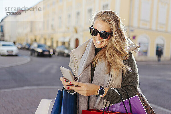 Smiling woman with shopping bags using smart phone on street