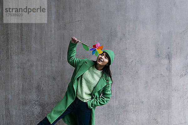 Cheerful woman playing with toy pinwheel in front of gray wall