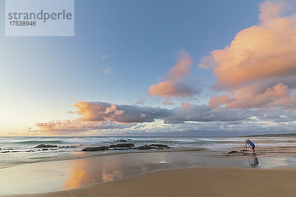 Male tourist taking photos on sandy coastal beach at moody sunrise