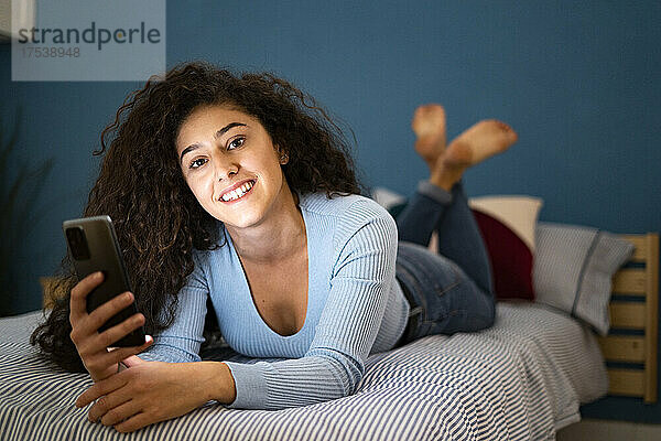 Young woman holding smart phone lying on bed