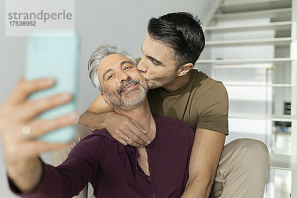 Happy gay man taking selfie with husband kissing on his cheek at home