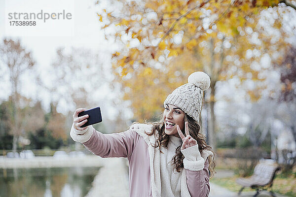 Glückliche Frau gestikuliert Friedenszeichen und macht im Park ein Selfie mit dem Handy