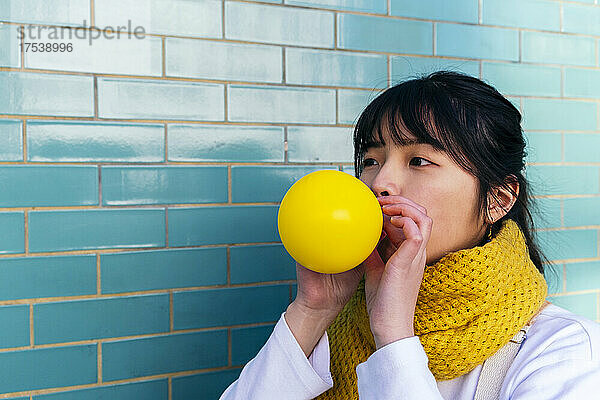 Woman blowing yellow balloon by turquoise brick wall