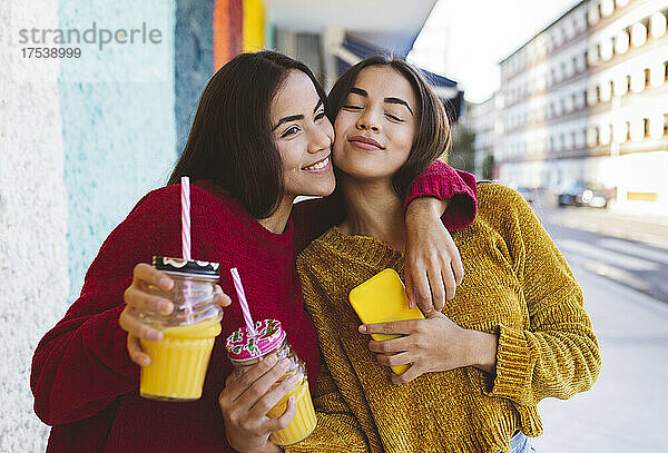 Smiling woman embracing twin sister with smart phone in city
