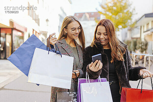 Glückliche Frauen mit Einkaufstüten  die ihr Smartphone auf dem Fußweg teilen