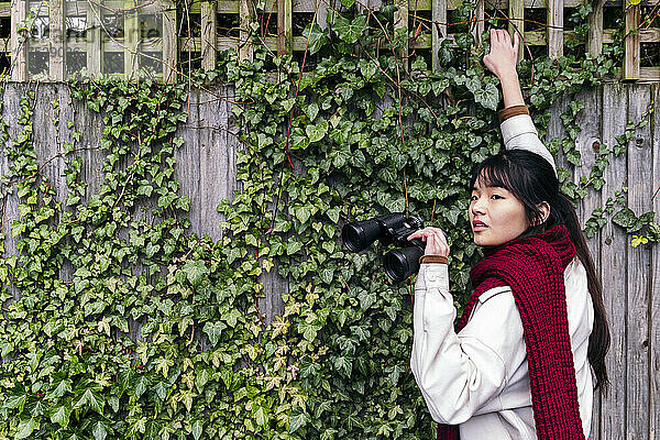 Young woman with binoculars standing near ivy wall