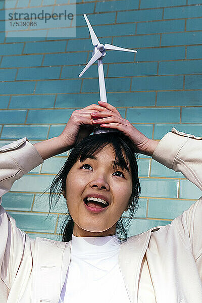 Playful young woman holding wind turbine toy on head in front of brick wall