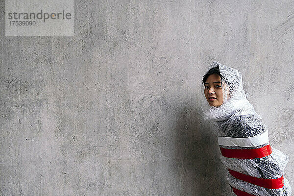Young woman protected in bubble wrap by gray wall