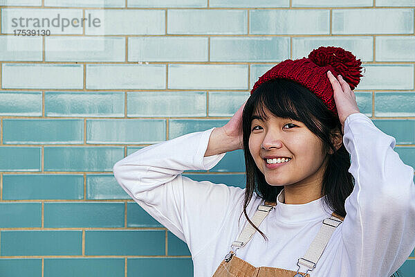 Smiling woman adjusting knit hat in front of turquoise wall