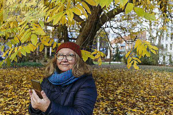 Lächelnde Seniorin mit Smartphone vor Herbstbaum im Park