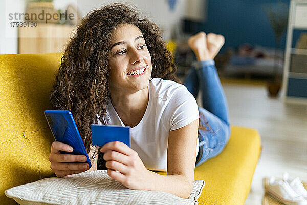 Smiling young woman with credit card and smart phone lying on sofa at home