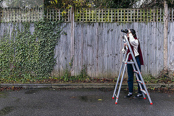 Young woman looking through binoculars standing by ladder on footpath