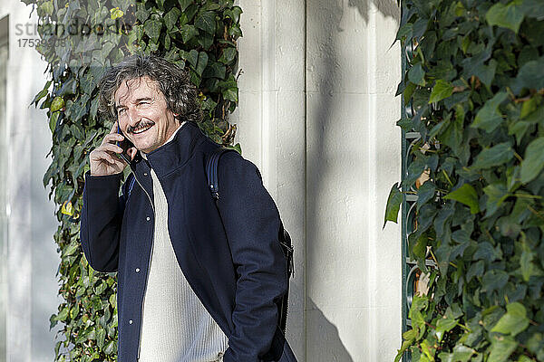 Lächelnder Mann  der an einem sonnigen Tag vor dem mit Efeublättern bedeckten Haus auf dem Smartphone spricht