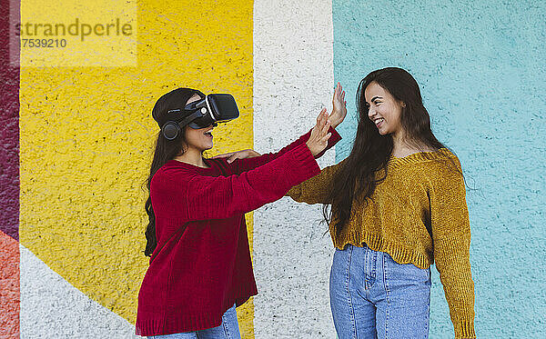 Smiling woman with sister wearing VR glasses gesturing by colorful wall