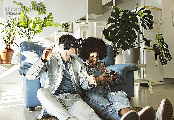 Junger Mann mit VR-Brille gestikuliert mit einer fröhlichen Freundin  die zu Hause ein Videospiel spielt