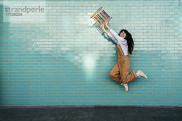 Excited woman jumping with abacus toy in front of turquoise wall