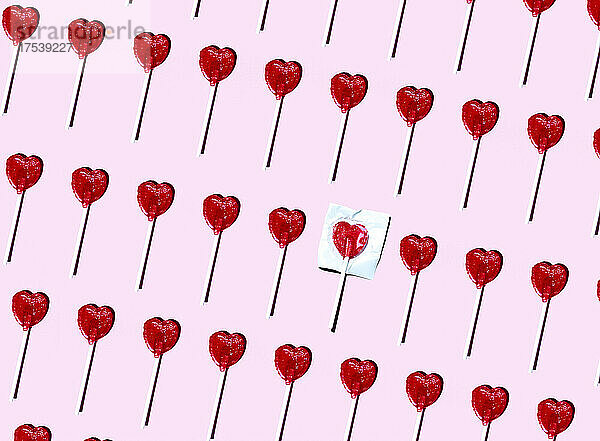 Pattern of heart shaped lollipops flat laid against pink background with single one still in wrapping paper